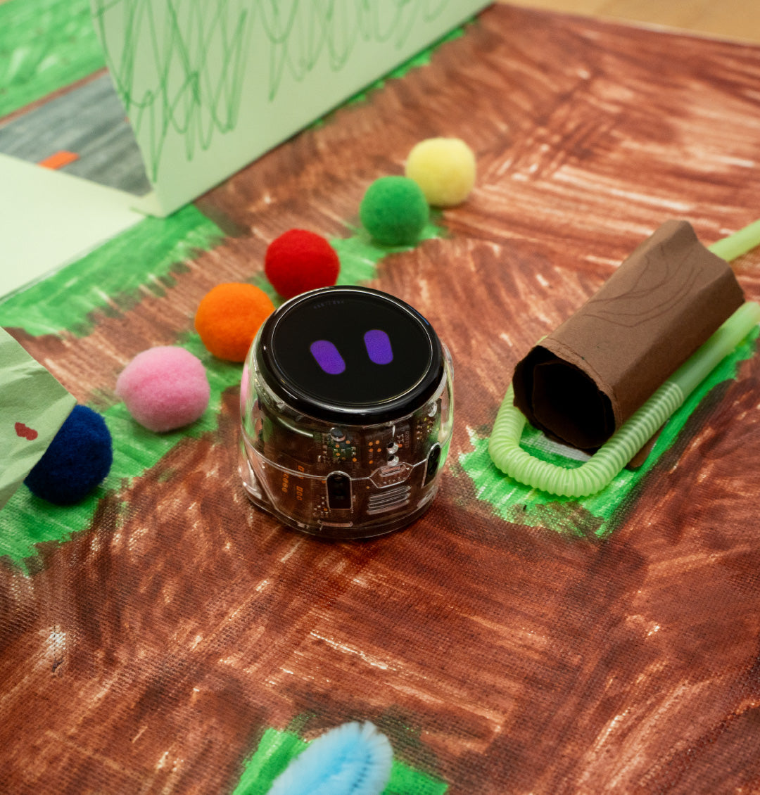 Ozobot Ari coding robot on a handmade nature-themed design mat featuring green spaces, pathways, and miniature landscape elements for a STEM project.
